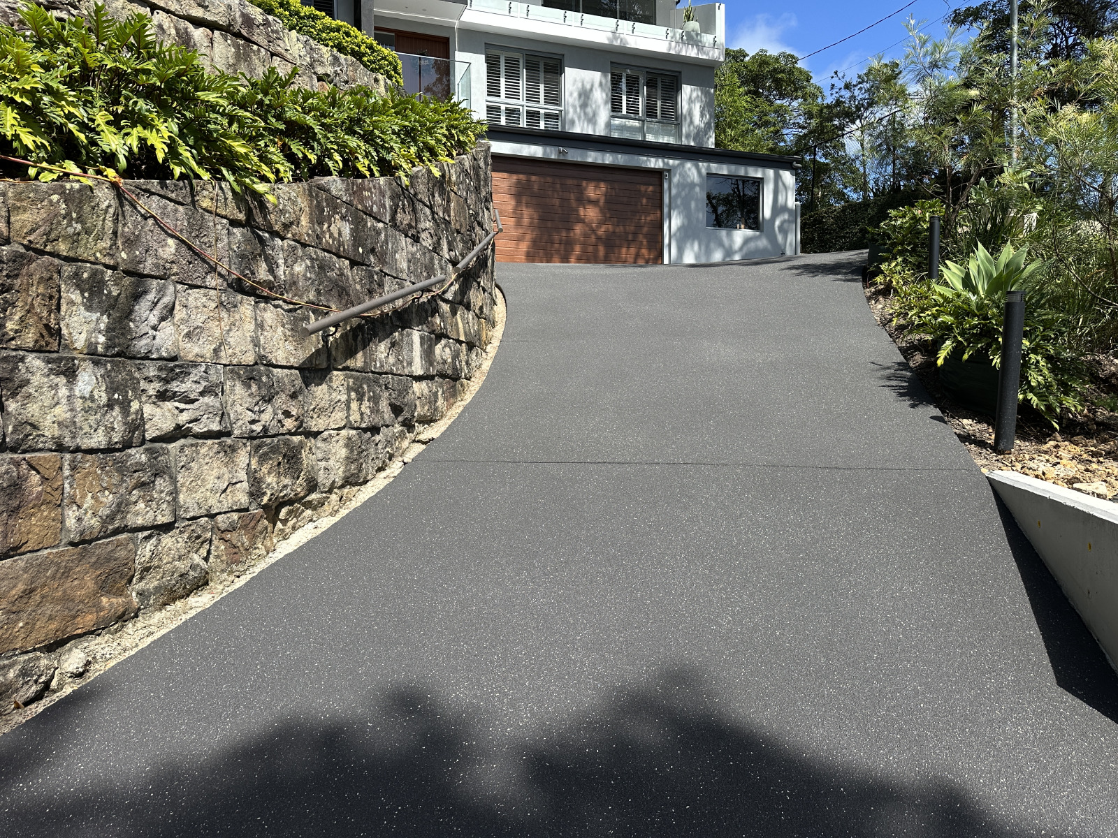 Steep curved driveway in Grays Point resurfaced in gunmetal grey with a white fleck anti-slip finish, leading up to a modern home’s timber garage door.